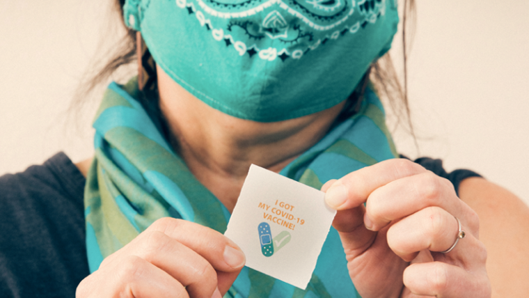 A Native elder shows off her vaccination sticker behind text: Be a Good Relative.